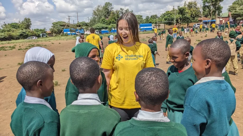 A participant in a yellow shirt engages with joyful children in green uniforms on a sunny school playground.